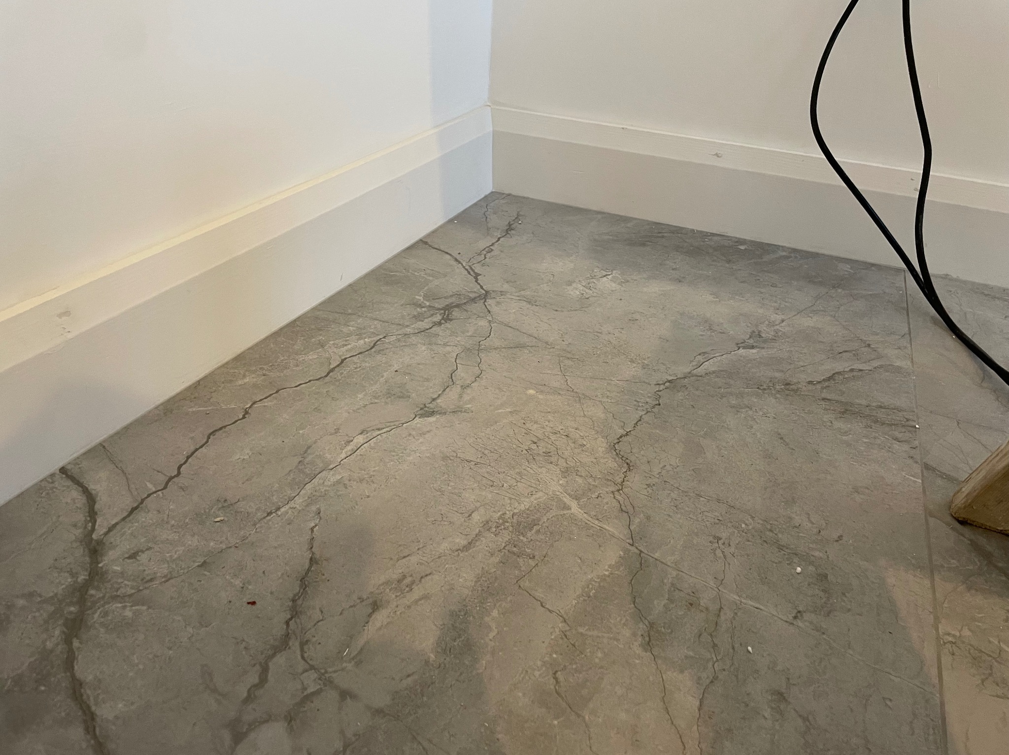 Szara podłoga z efektem marmuru, widoczne białe listwy przypodłogowe i fragment kabla elektrycznego w rogu pomieszczenia.