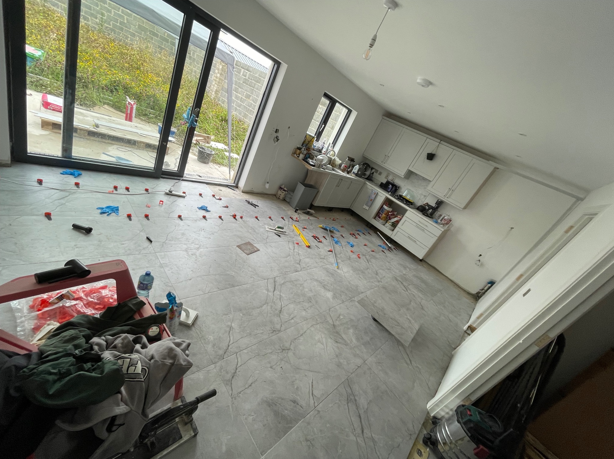 Wnętrze w trakcie prac: szare płytki podłogowe z marmurkowym wzorem, poziomica, krzyżyki dystansowe, widok na okno tarasowe i zabudowę kuchenną w tle.