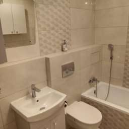 Krzysztof Jarzmik - Usługi remontowo-budowlane - Jasna łazienka po remoncie z umywalką na szafce, wiszącą toaletą i wanną z prysznicem, wykończona płytkami w odcieniach beżu z dekoracyjnym wzorem.