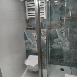 Krzysztof Jarzmik - Usługi remontowo-budowlane - Nowoczesna łazienka z kabiną prysznicową ze szklanymi drzwiami, toaleta z folią ochronną, grzejnik drabinkowy i płytki imitujące marmur w odcieniach bieli i turkusu.