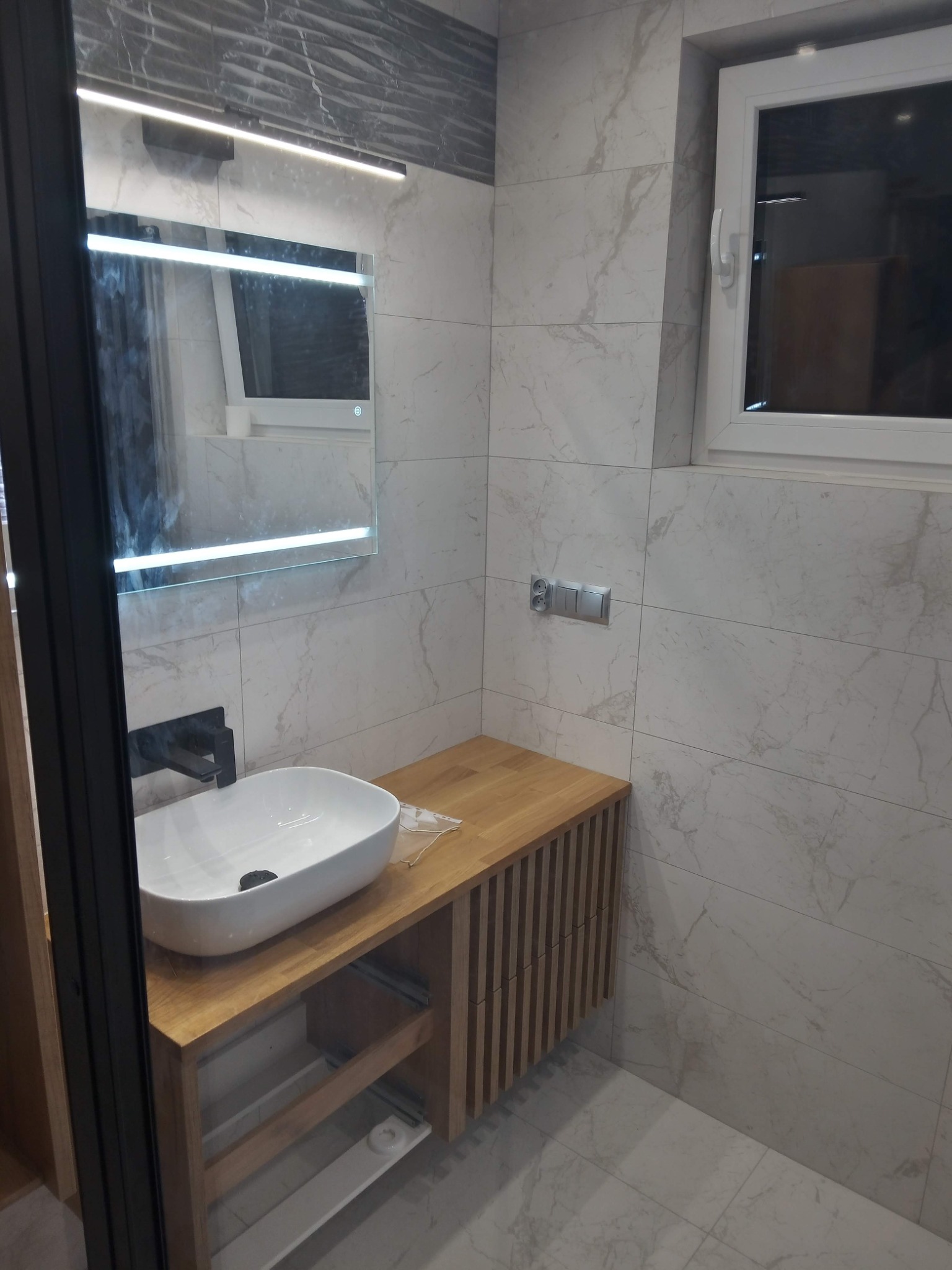 Nowoczesna łazienka z marmurowymi płytkami, drewnianą szafką pod umywalką i podświetlanym lustrem LED.