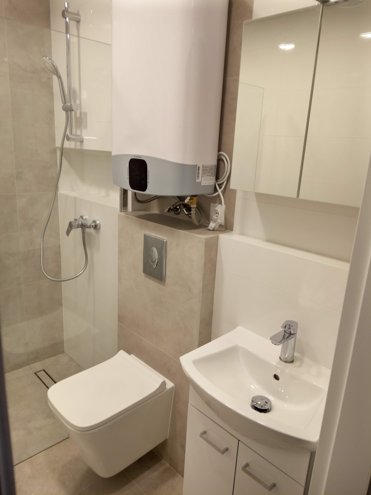 Nowoczesna, jasna łazienka z podwieszaną toaletą, umywalką z szafką, kabiną prysznicową typu walk-in i podgrzewaczem wody marki Ariston