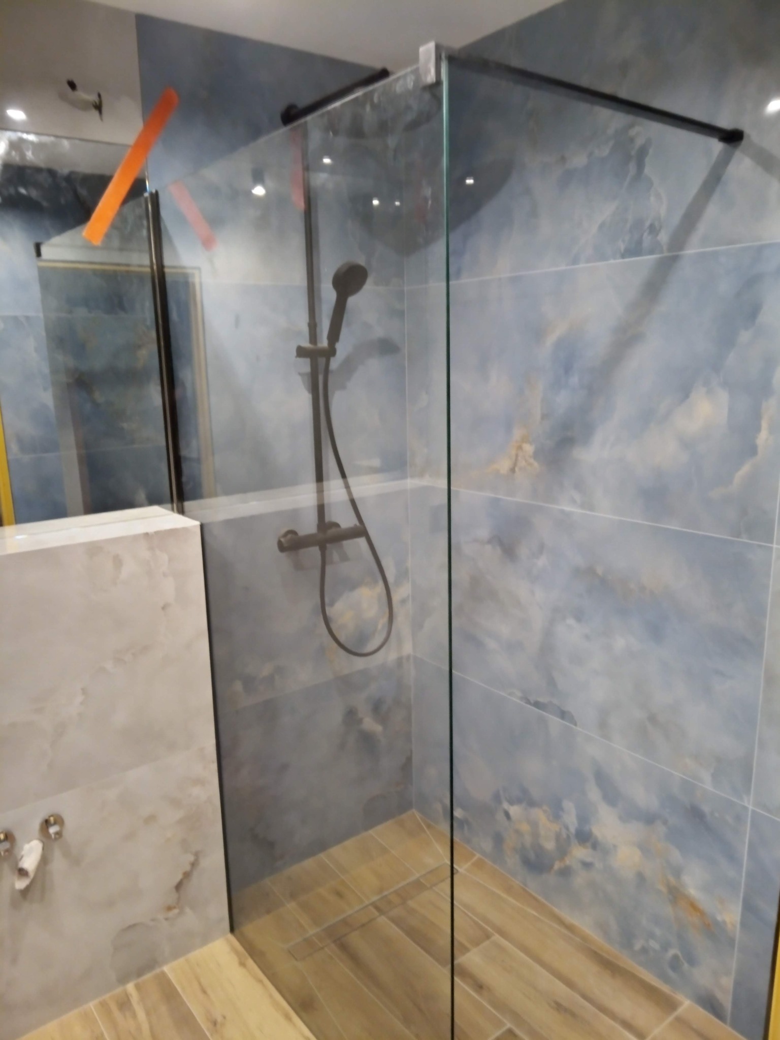 Szklana kabina prysznicowa z deszczownicą, wykończona płytkami imitującymi marmur w odcieniach błękitu i beżu, z drewnopodobną podłogą i chromowanymi elementami armatury.