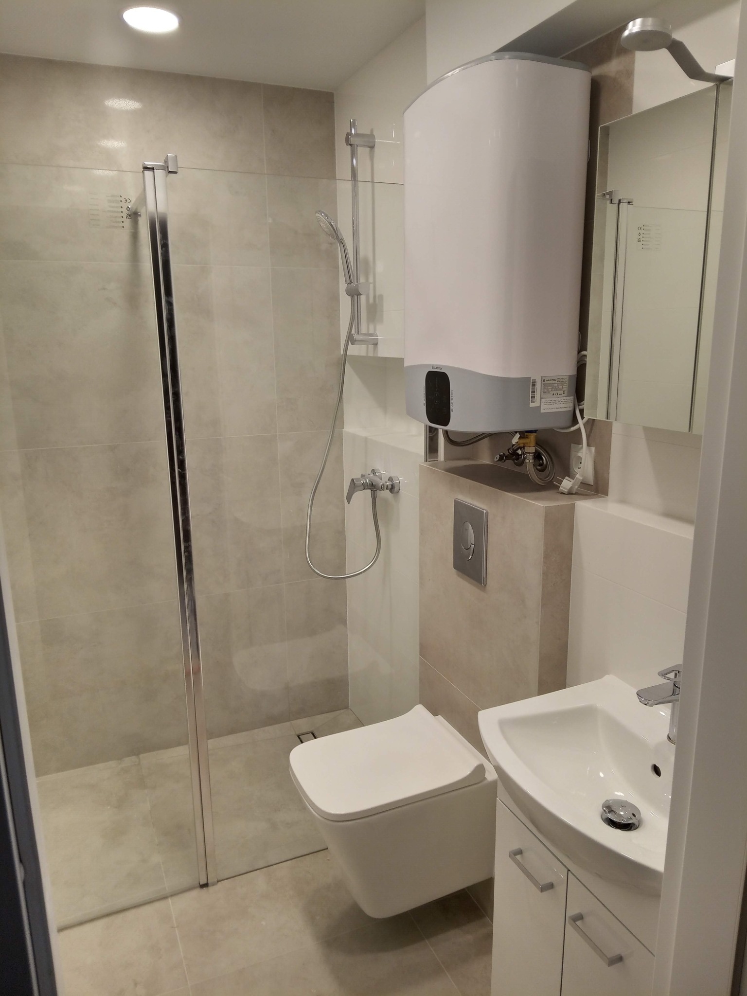 Nowoczesna łazienka z kabiną prysznicową ze szklanymi drzwiami, podwieszaną toaletą, umywalką z szafką i bojlerem elektrycznym.