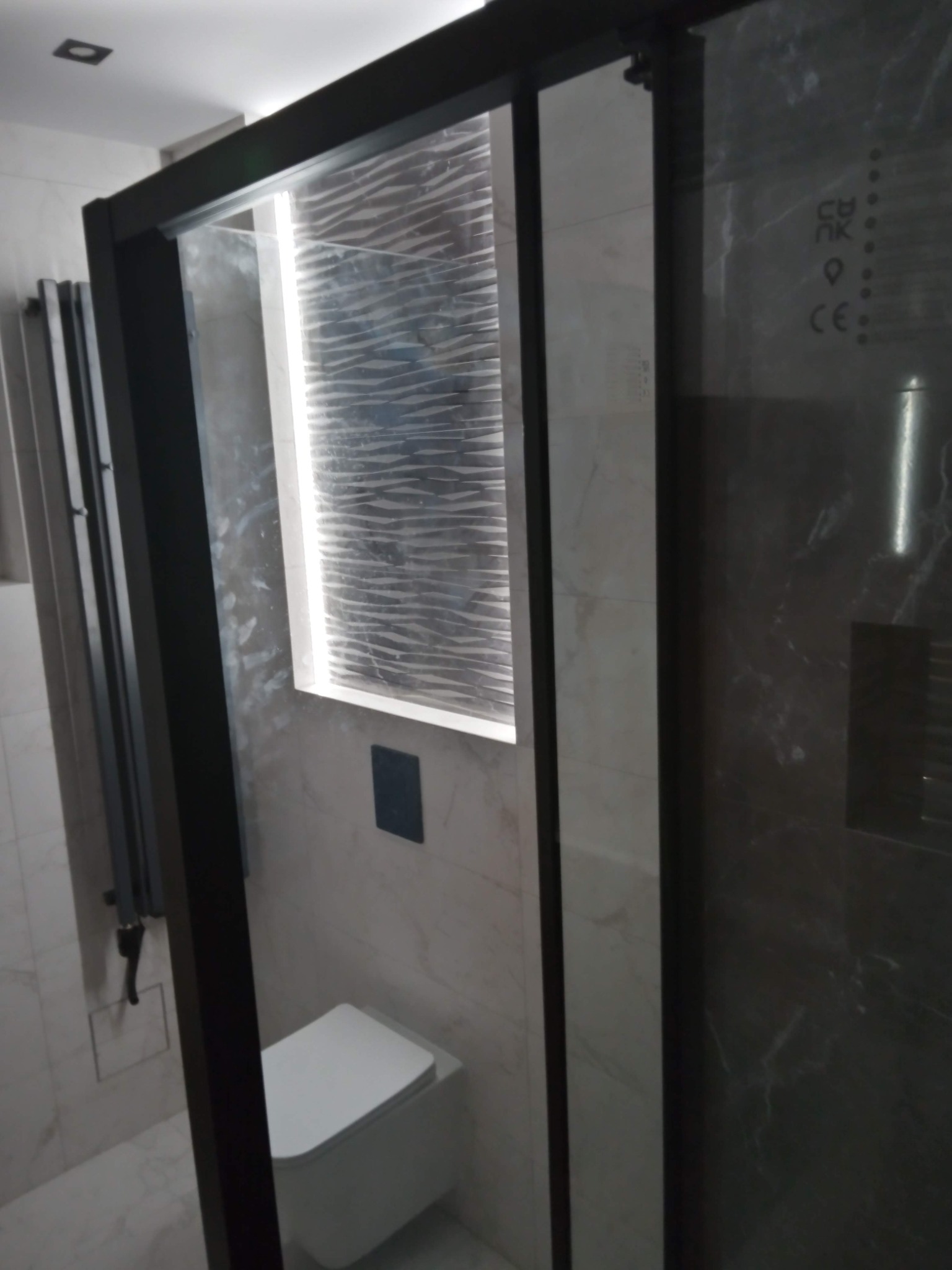 Nowoczesna kabina prysznicowa z czarną ramą i wzorzystą ścianą wewnątrz, widoczna toaleta podwieszana i grzejnik drabinkowy, wykończenie marmurowe