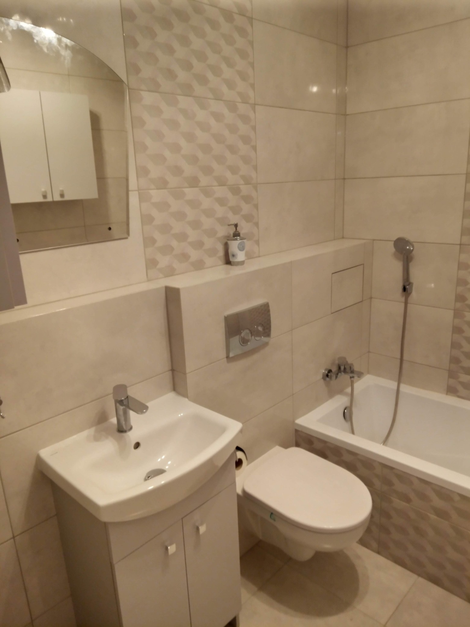 Jasna łazienka po remoncie z umywalką na szafce, wiszącą toaletą i wanną z prysznicem, wykończona płytkami w odcieniach beżu z dekoracyjnym wzorem.