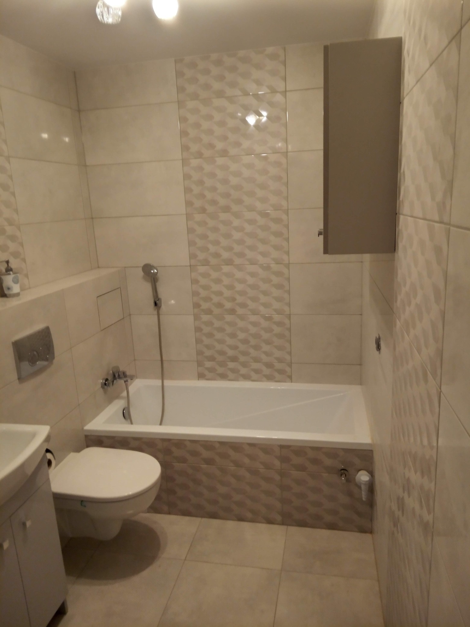 Wykończona łazienka z beżowymi i białymi płytkami o wzorze 3D, wisząca szafka nad wanną, toaleta podwieszana i umywalka z szafką w rogu.