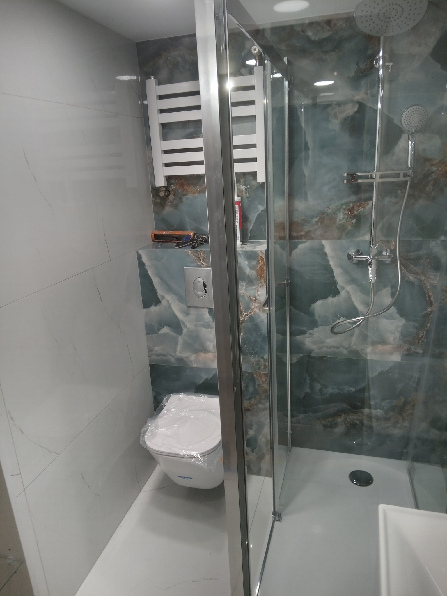 Nowoczesna łazienka z kabiną prysznicową ze szklanymi drzwiami, toaleta z folią ochronną, grzejnik drabinkowy i płytki imitujące marmur w odcieniach bieli i turkusu.