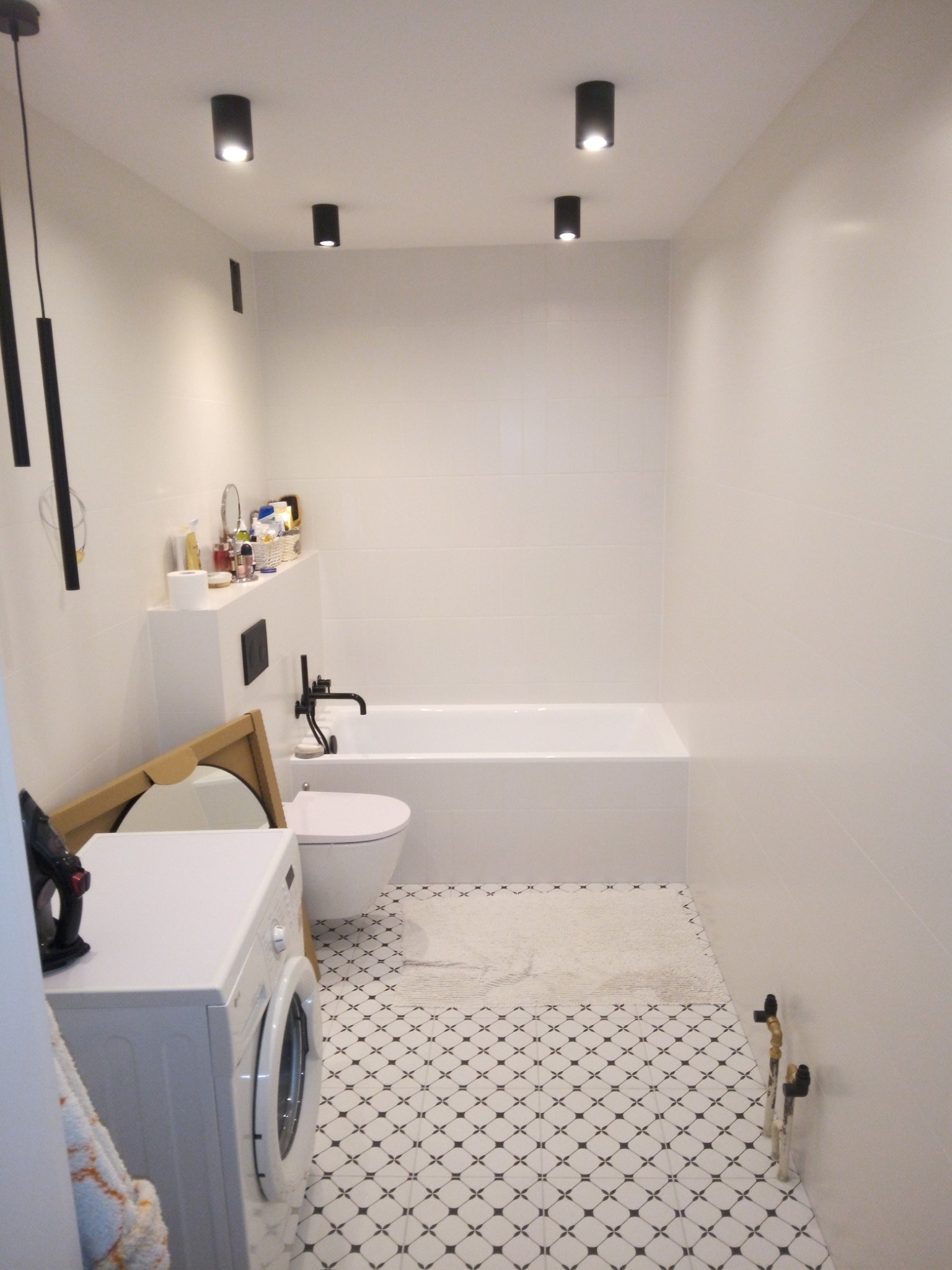 Wykończona, jasna łazienka w Nowym Sączu z białą armaturą, czarnymi punktowymi lampami sufitowymi i wzorzystą podłogą. W rogu widoczna pralka i rozpakowane lustro.