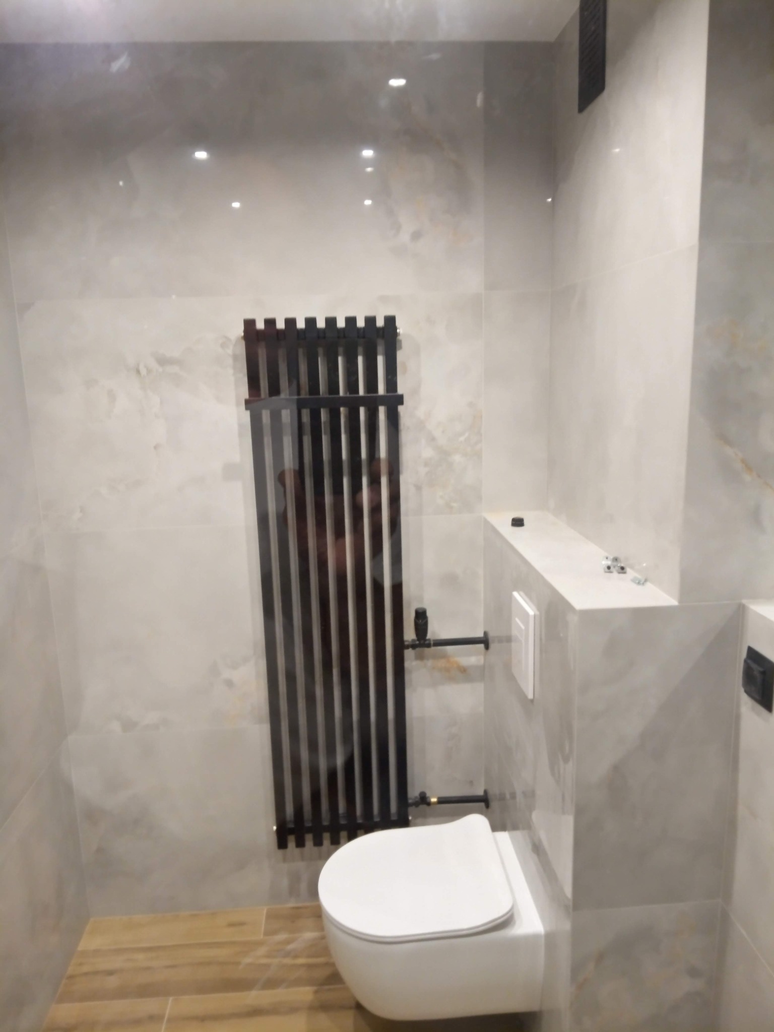 Nowoczesna łazienka z wiszącą toaletą, grzejnikiem drabinkowym w kolorze antracytu i płytkami imitującymi marmur na ścianach oraz drewnopodobną podłogą.