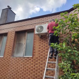Anemos Bartosz Naskręt - Mężczyzna na drabinie montuje klimatyzator na elewacji budynku z cegły klinkierowej, widoczny fragment drzewa i komin.