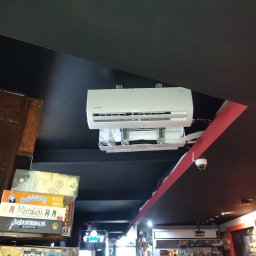 Anemos Bartosz Naskręt - Biała klimatyzacja marki Rotenso zamontowana pod ciemnym sufitem w lokalu komercyjnym, widoczne gry planszowe na półce w tle.