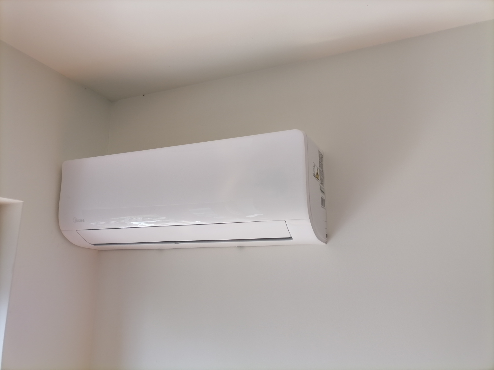 Biała jednostka wewnętrzna klimatyzacji Midea zamontowana na jasnej ścianie w rogu pomieszczenia.