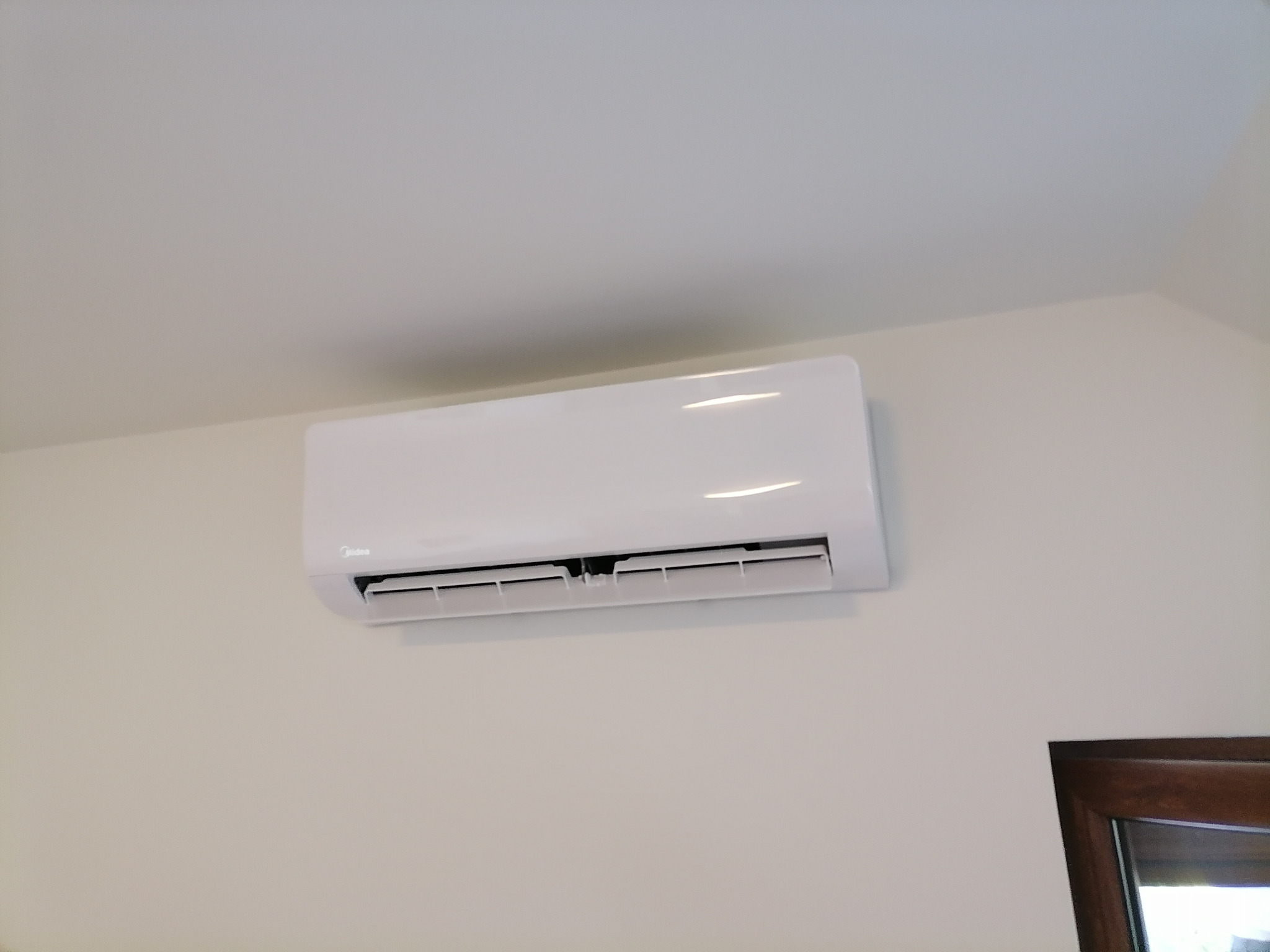 Biały klimatyzator Midea zamontowany na jasnej ścianie, widoczny fragment ciemnej ramy okiennej w prawym dolnym rogu.