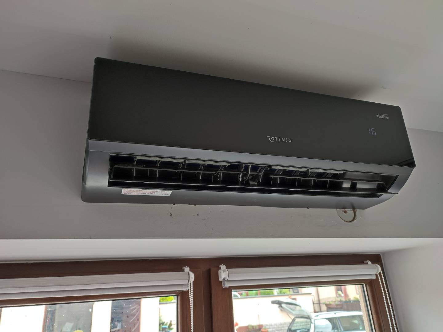 Czarny klimatyzator Rotenso zamontowany na ścianie w pomieszczeniu z oknem, widoczny wyświetlacz temperatury ustawionej na 16 stopni, poniżej okno z roletami.