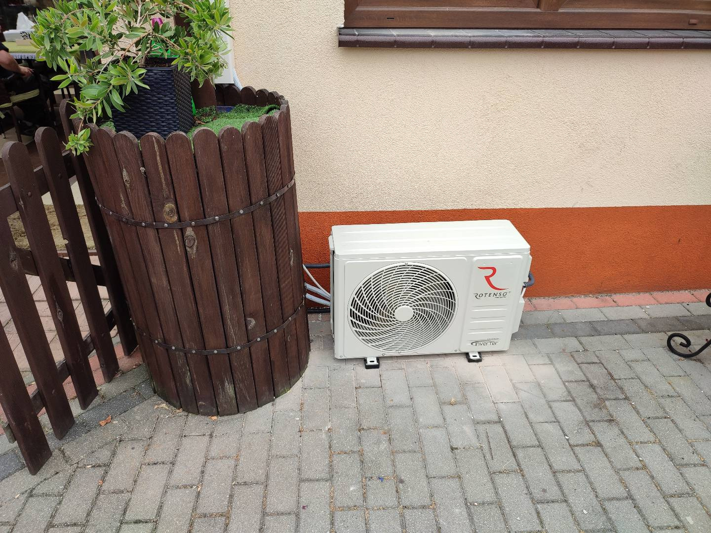Zewnętrzna jednostka klimatyzacji Rotenso umieszczona na brukowanym chodniku obok drewnianej donicy z roślinami, na tle ściany budynku z brązowym pasem.
