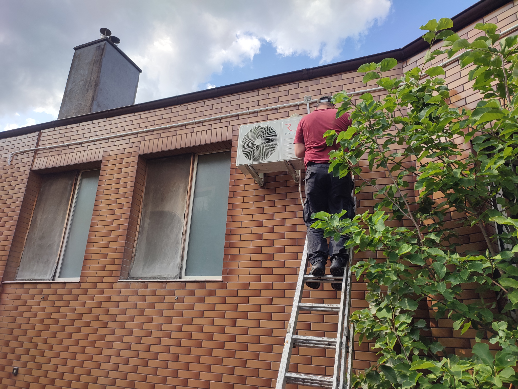 Mężczyzna na drabinie montuje klimatyzator na elewacji budynku z cegły klinkierowej, widoczny fragment drzewa i komin.