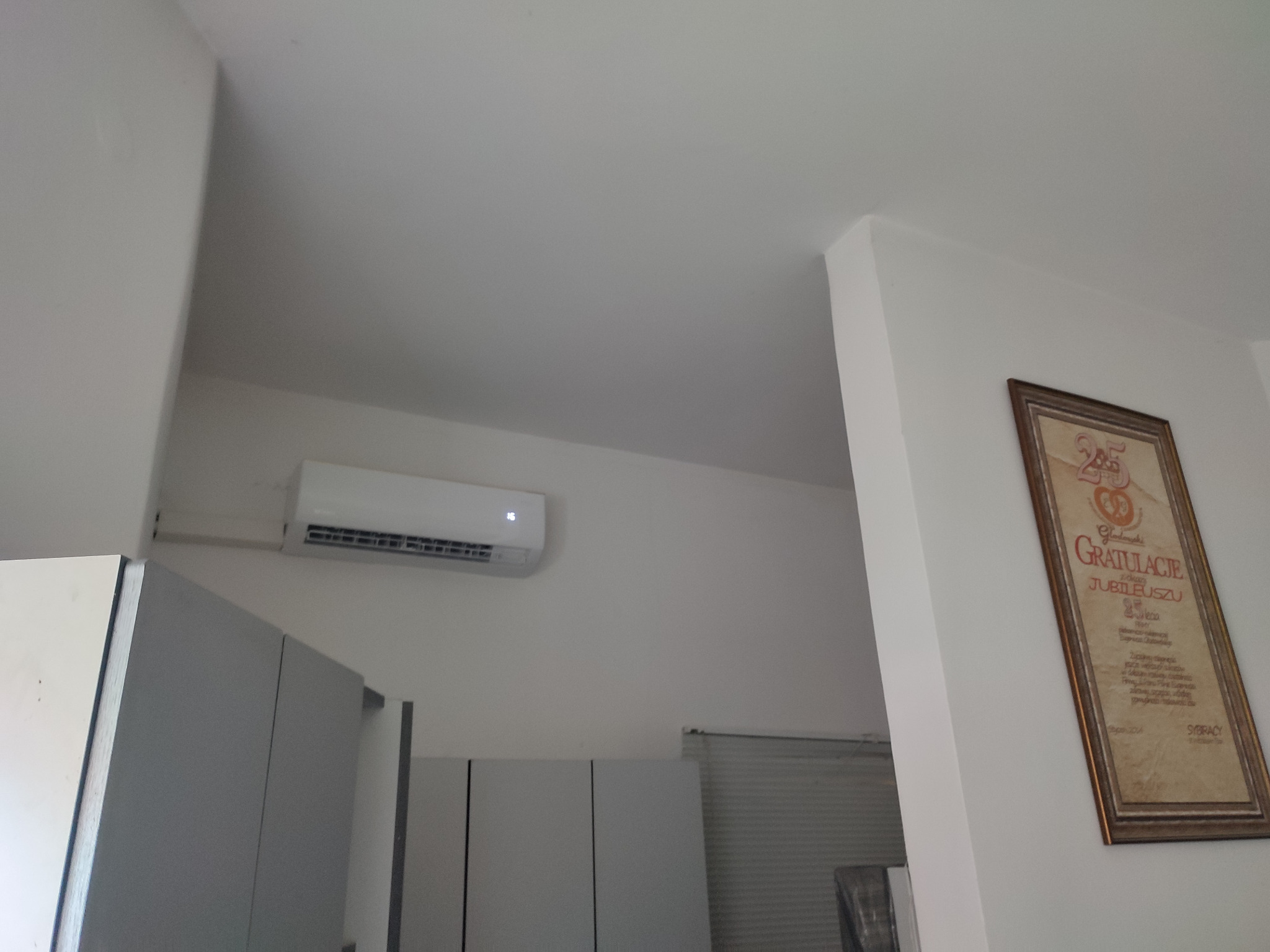 Biały klimatyzator ścienny z wyświetlaczem temperatury zamontowany w pomieszczeniu, widoczna część zabudowy meblowej i oprawiony dyplom na ścianie.