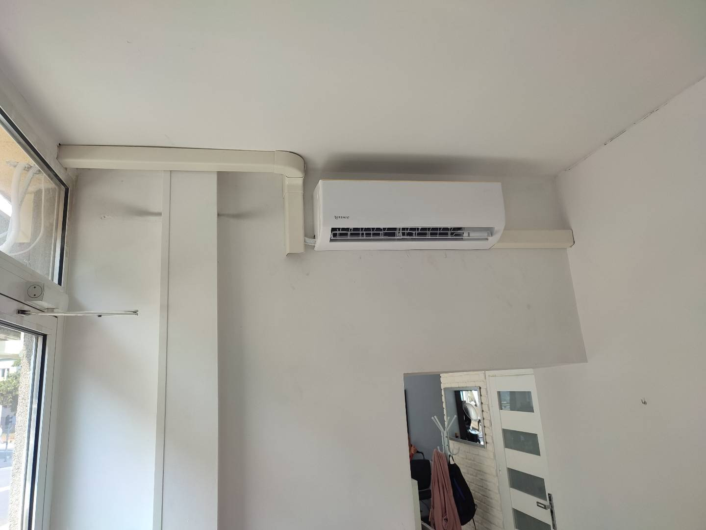 Biała jednostka klimatyzacji marki Rotenic zamontowana na ścianie w pomieszczeniu z widocznymi elementami instalacji elektrycznej i fragmentem okna balkonowego.