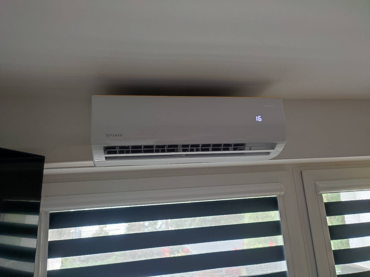 Biała klimatyzacja Rotenso Roni, zamontowana nad oknem z żaluzjami poziomymi, wyświetlająca temperaturę 16 stopni Celsjusza.