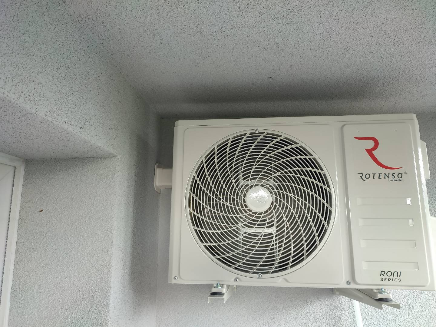 Biała jednostka zewnętrzna klimatyzacji Rotenso Roni Series zamontowana na szarej, chropowatej ścianie budynku, widok od dołu.