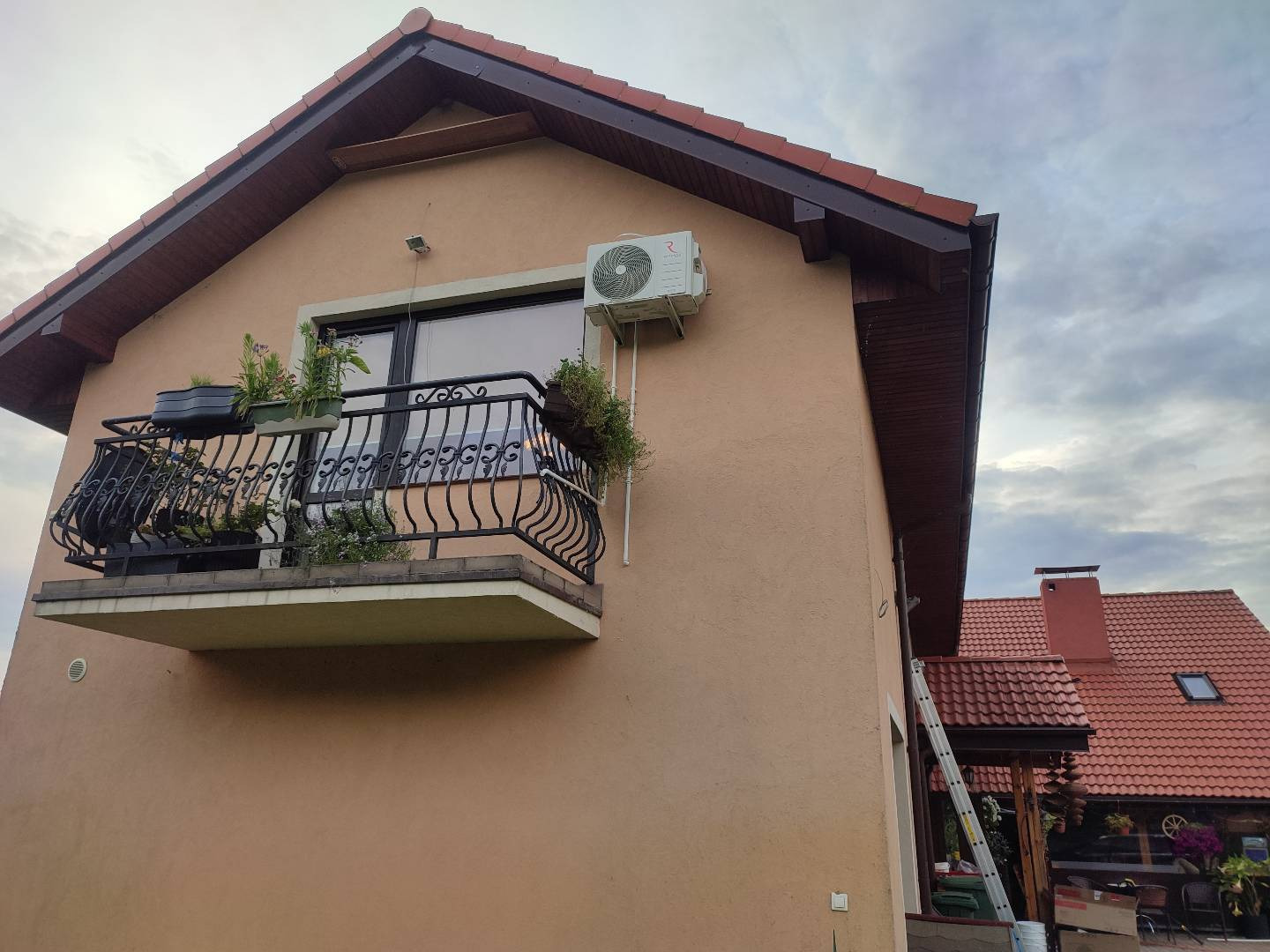 Zewnętrzna jednostka klimatyzacji zamontowana na ścianie domu z balkonem i roślinami w doniczkach. Widoczna drabina oparta o ścianę budynku.