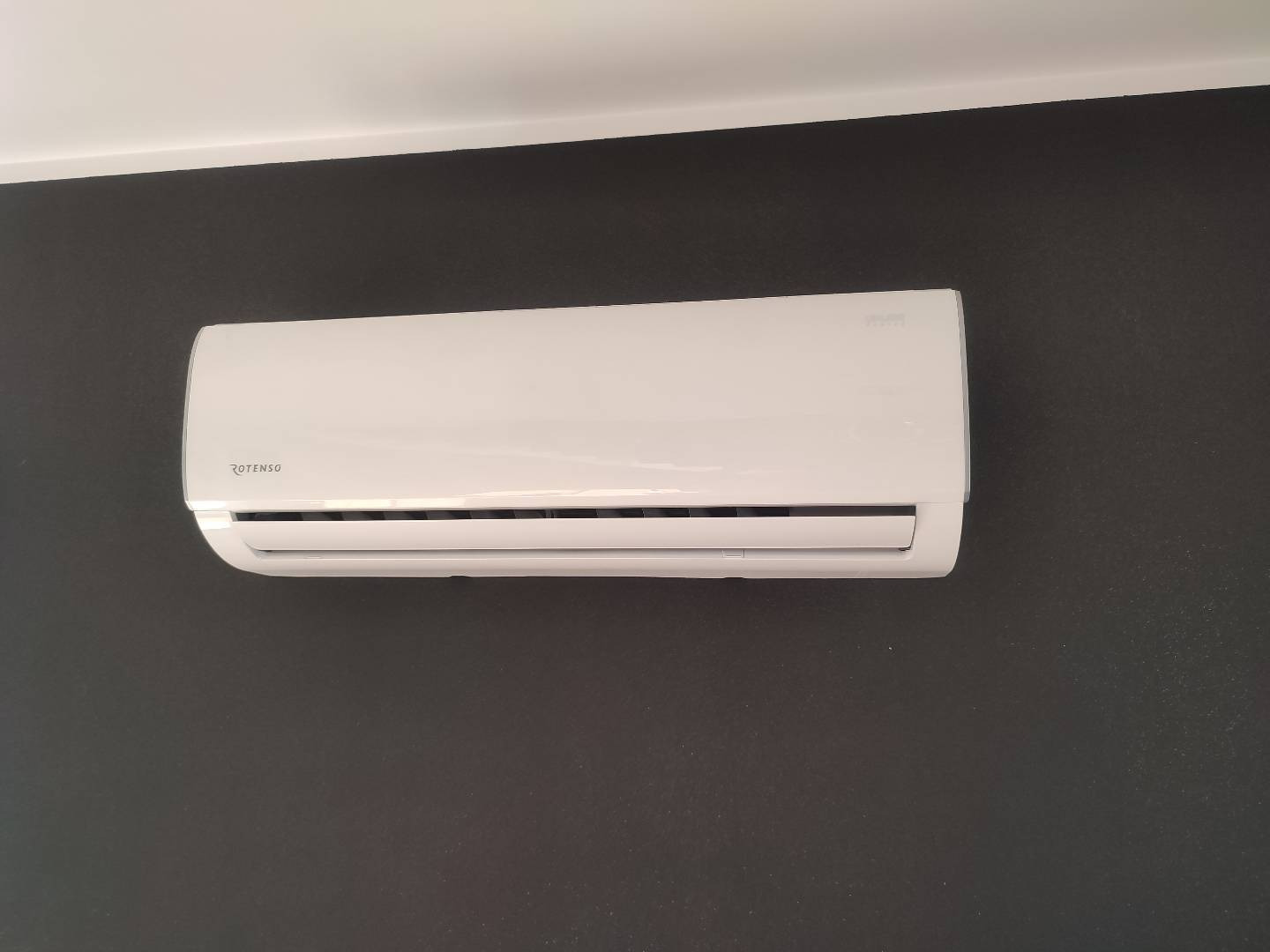 Biała jednostka klimatyzacji Rotenso zamontowana na ciemnoszarej ścianie, widok z przodu.
