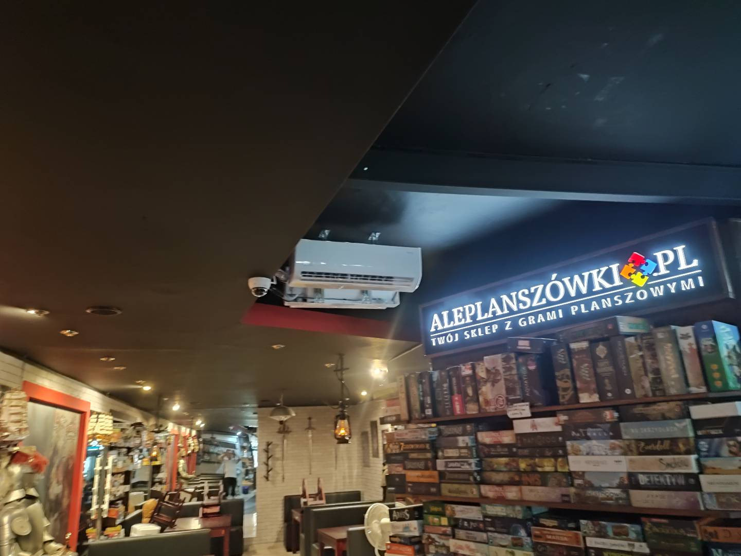 Wnętrze sklepu z grami planszowymi: widoczna klimatyzacja kasetonowa zamontowana pod sufitem, regały wypełnione grami, oświetlenie punktowe i dekoracyjne lampy wiszące.