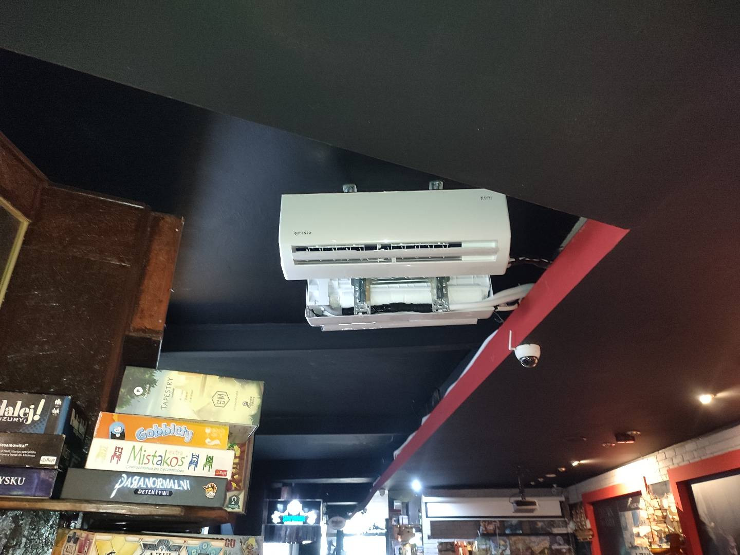 Biała klimatyzacja marki Rotenso zamontowana pod ciemnym sufitem w lokalu komercyjnym, widoczne gry planszowe na półce w tle.