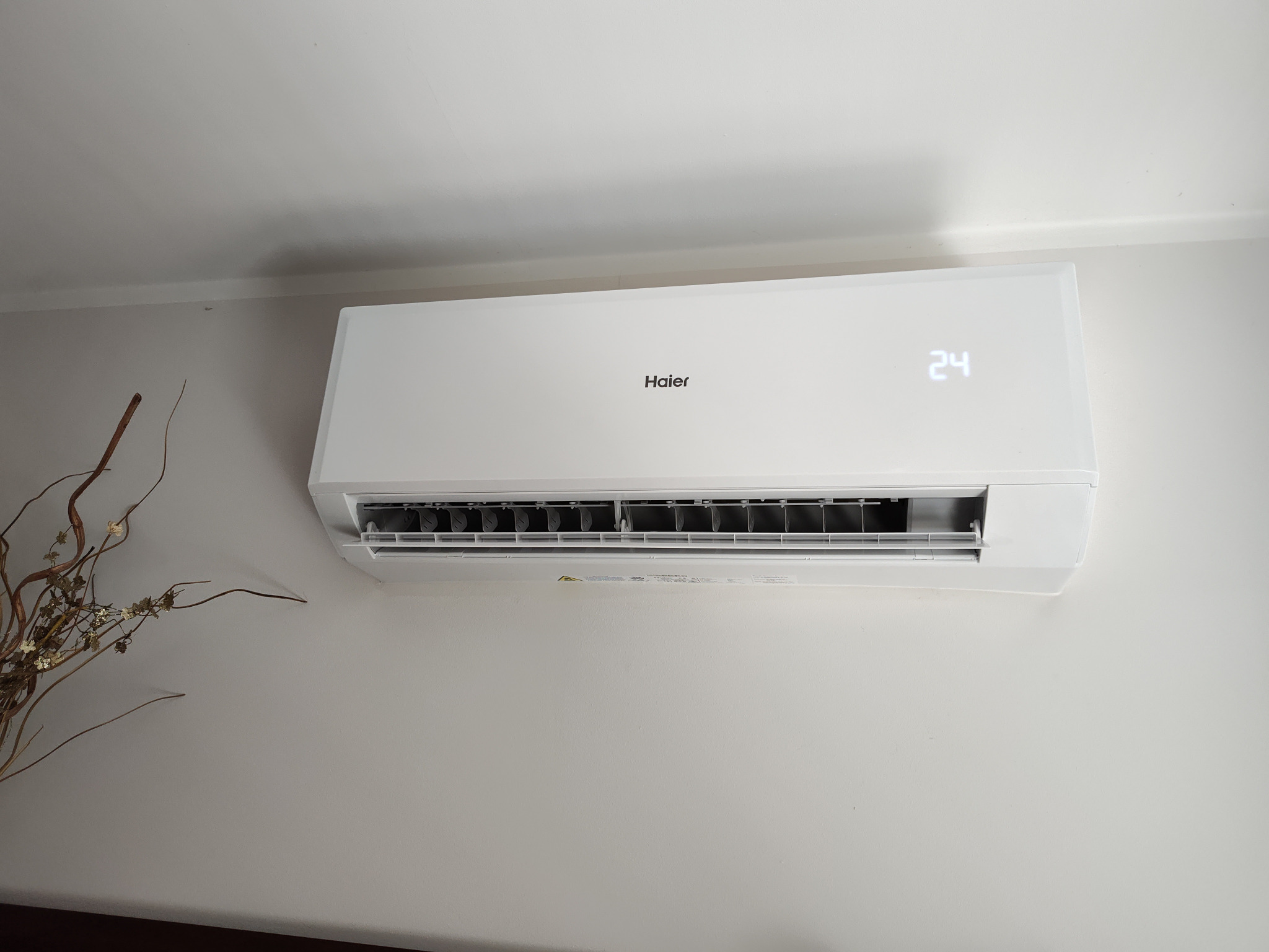 Biała klimatyzacja Haier zamontowana na ścianie, wyświetlająca temperaturę 24 stopnie, z dekoracyjnymi gałązkami po lewej stronie.