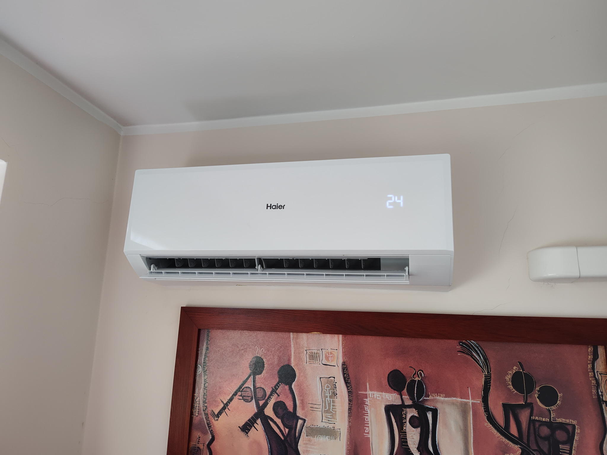 Biała klimatyzacja Haier zamontowana na ścianie nad obrazem w brązowej ramie, wyświetlająca temperaturę 24 stopnie.