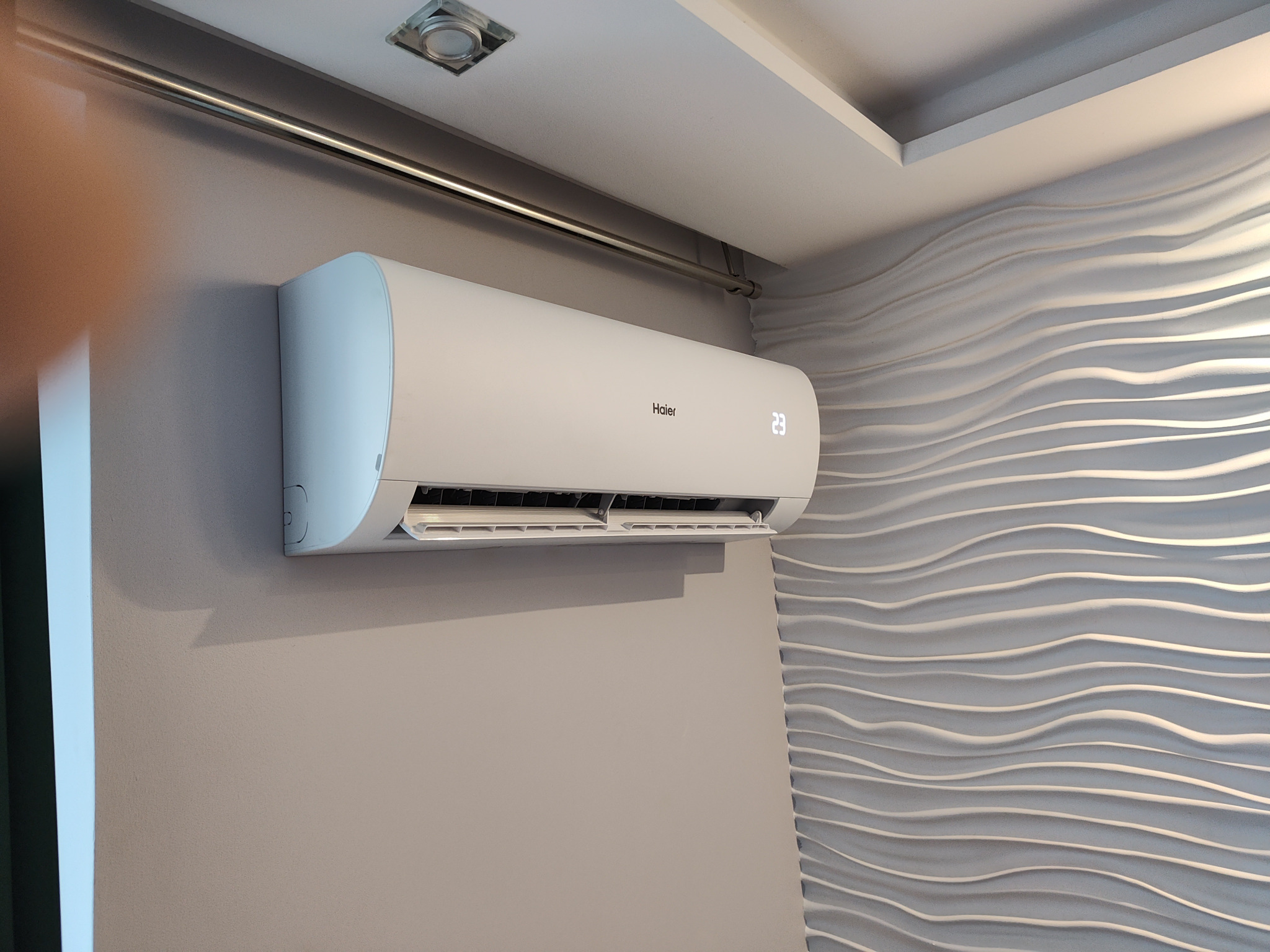 Biała klimatyzacja Haier zamontowana na szarej ścianie, wyświetlająca temperaturę 23 stopnie, widoczna dekoracyjna ściana w tle.
