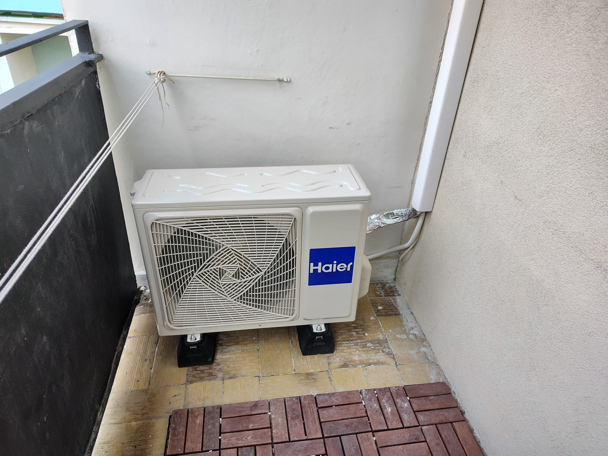 Kompaktowa jednostka zewnętrzna klimatyzatora Haier, zamontowana na czarnych podkładkach antywibracyjnych na balkonie z drewnianą podłogą, widoczne przewody i fragment ściany budynku.