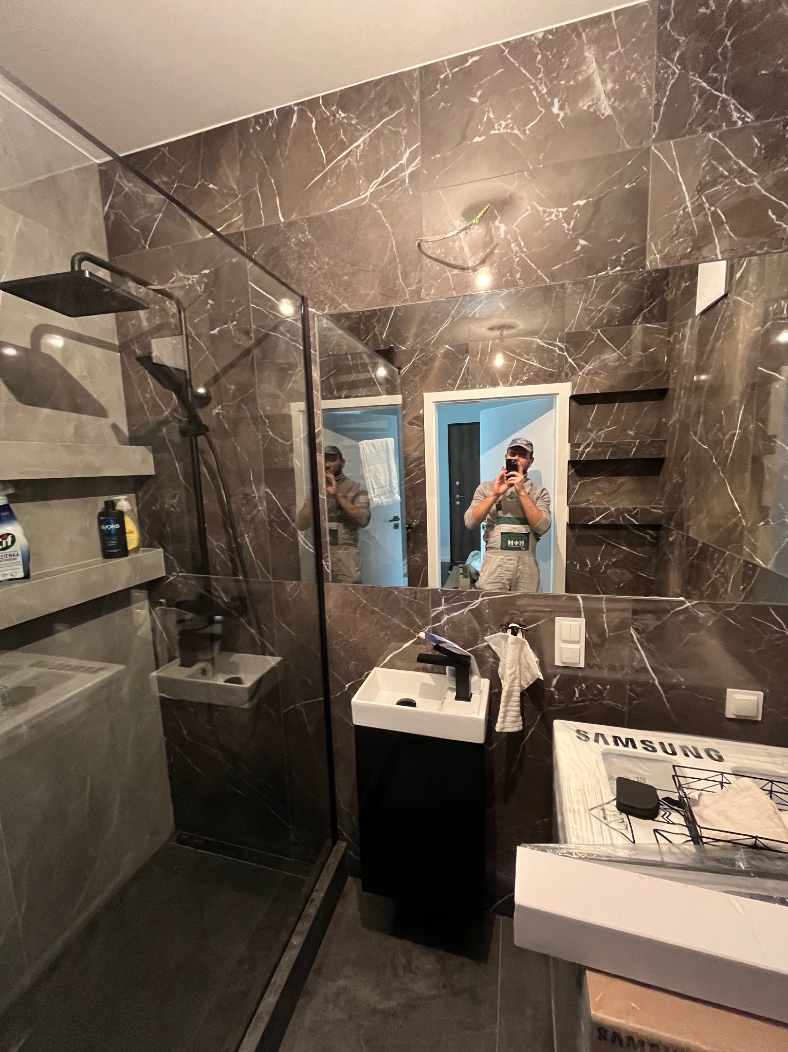 Nowoczesna łazienka z kabiną prysznicową typu walk-in, wykończona płytkami imitującymi marmur, z czarną armaturą i szafką pod umywalką. W lustrze odbija się osoba robiąca zdjęcie.