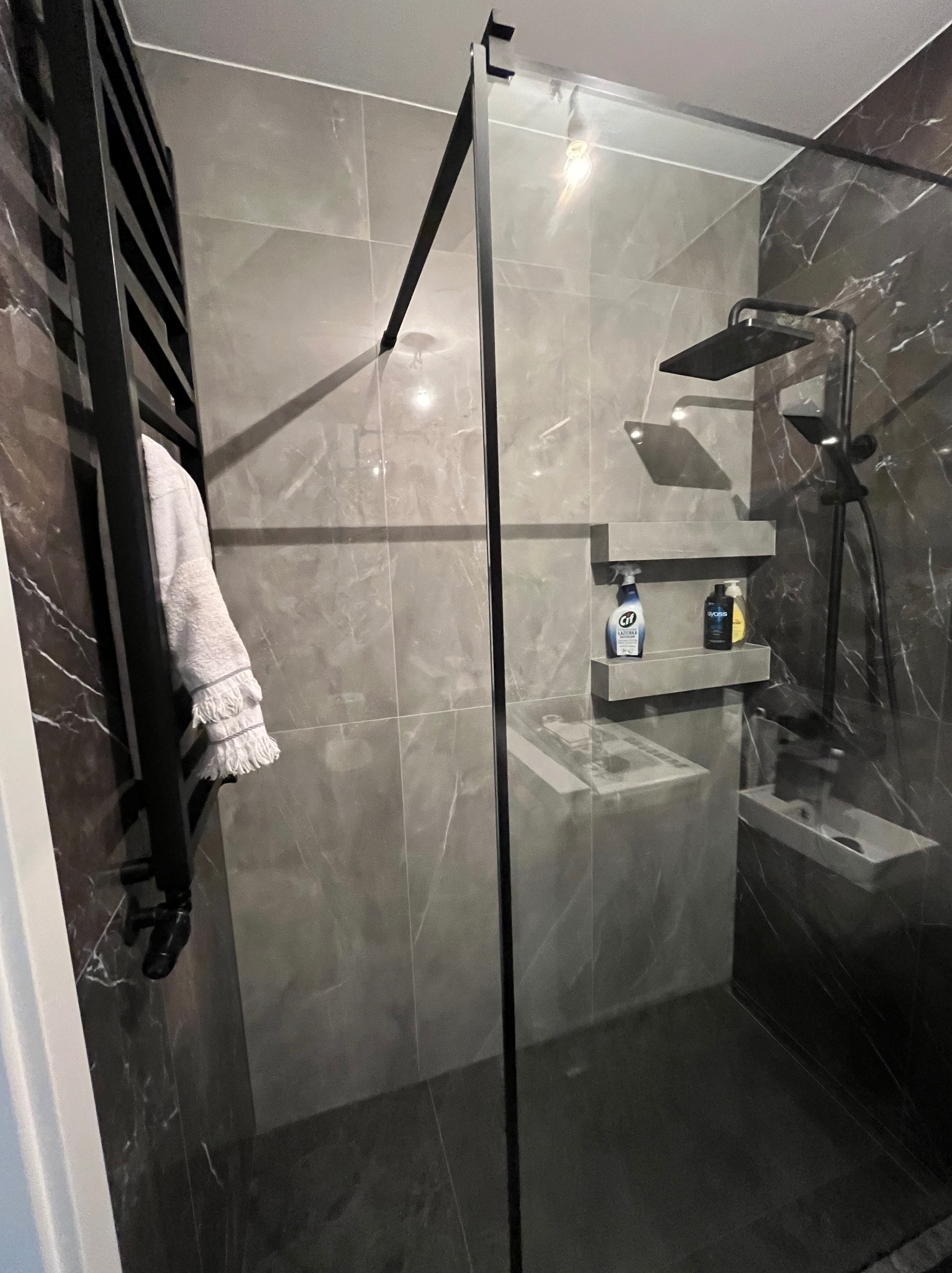 Nowoczesna kabina prysznicowa walk-in z czarnymi profilami i deszczownicą, marmurowe płytki w odcieniach szarości, grzejnik drabinkowy z ręcznikiem i półki z kosmetykami.
