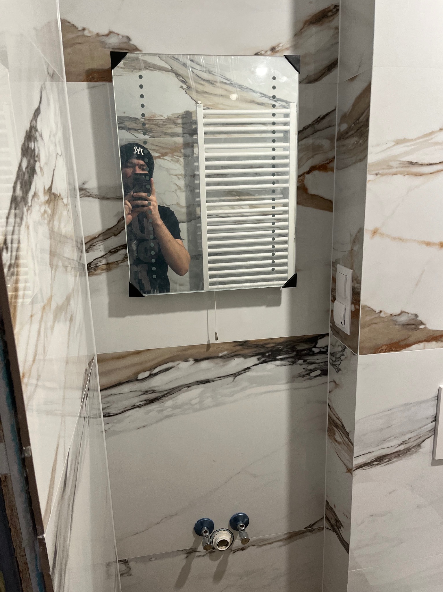 Wykończona łazienka z marmurowymi płytkami, widoczne przyłącza wodne, grzejnik drabinkowy w lustrze z oświetleniem LED, odbicie fotografa w czapce.