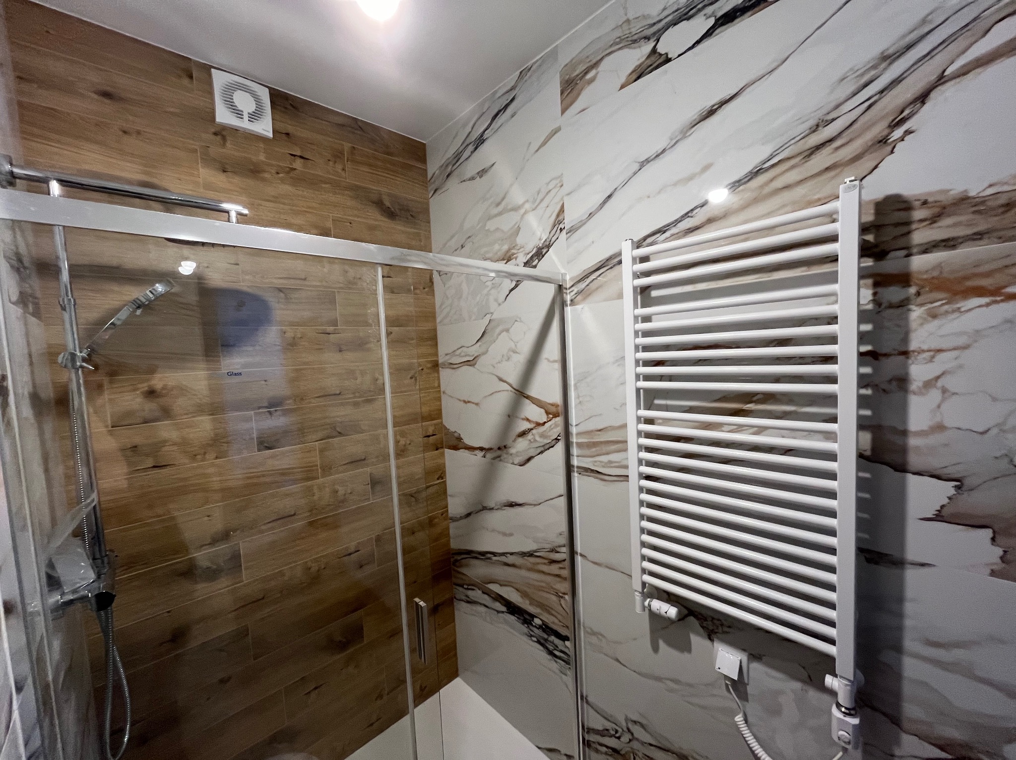 Kabina prysznicowa z chromowanymi profilami i drewnopodobnymi płytkami na ścianie, obok grzejnik drabinkowy na ścianie z marmurowym wzorem.