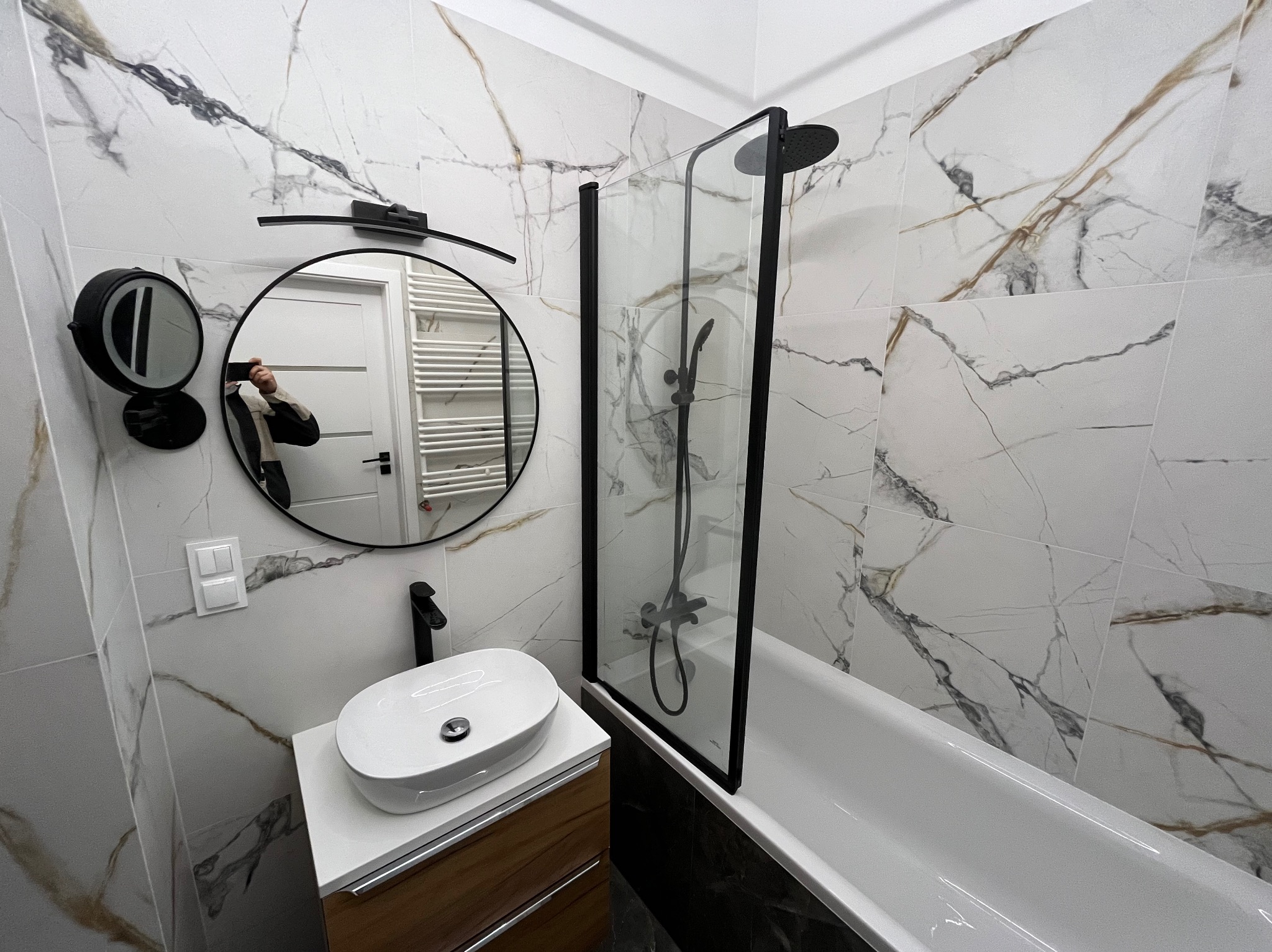 Nowoczesna łazienka z białymi marmurkowymi płytkami, okrągłym lustrem z podświetleniem, białą umywalką na drewnianej szafce i czarną armaturą, kabina prysznicowa z czarną ramą przy wannie.
