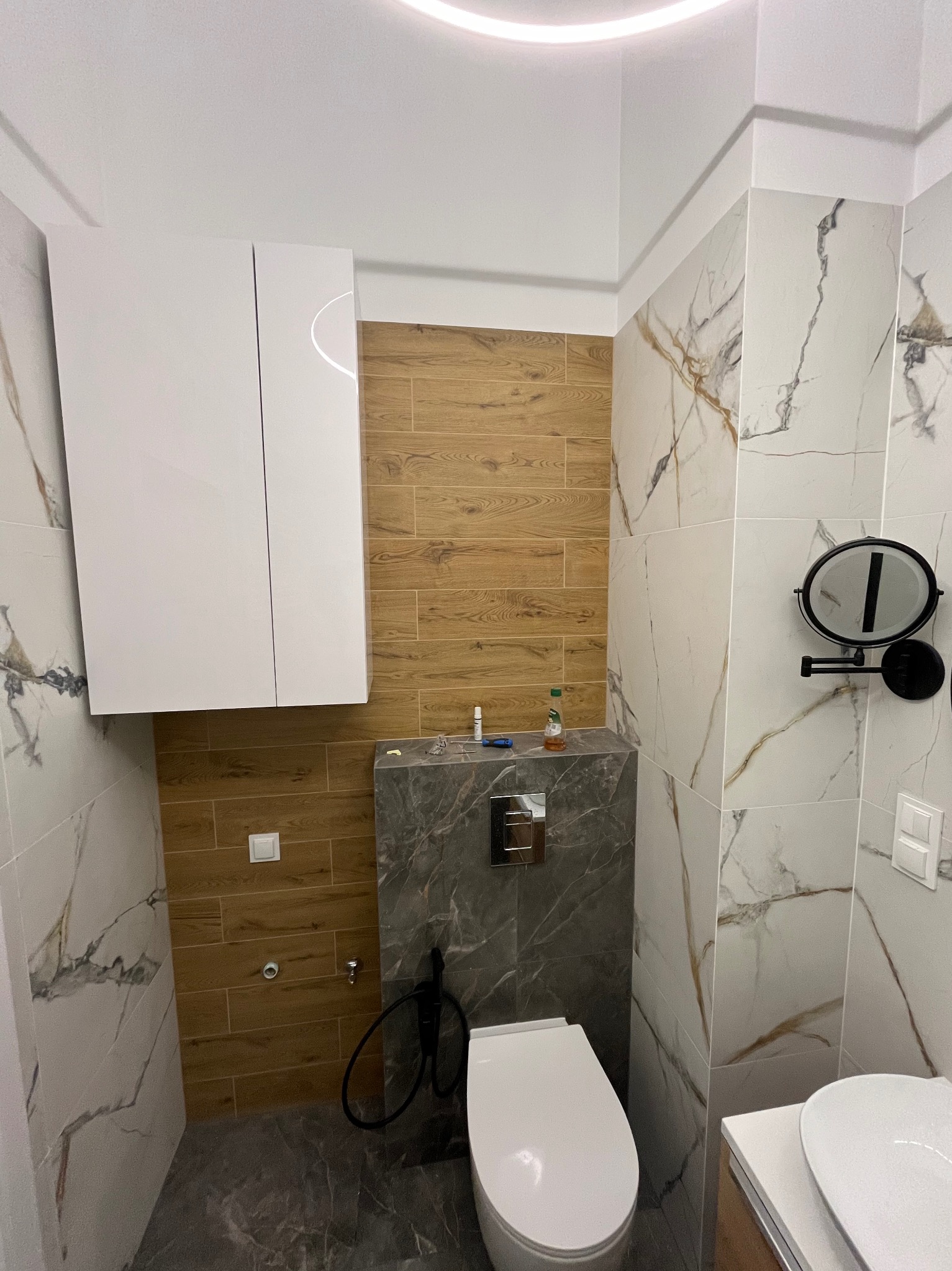 Nowoczesna łazienka po renowacji: połączenie płytek imitujących drewno i marmur, podwieszana toaleta z szarą obudową, biała szafka wisząca i okrągłe lustro kosmetyczne.