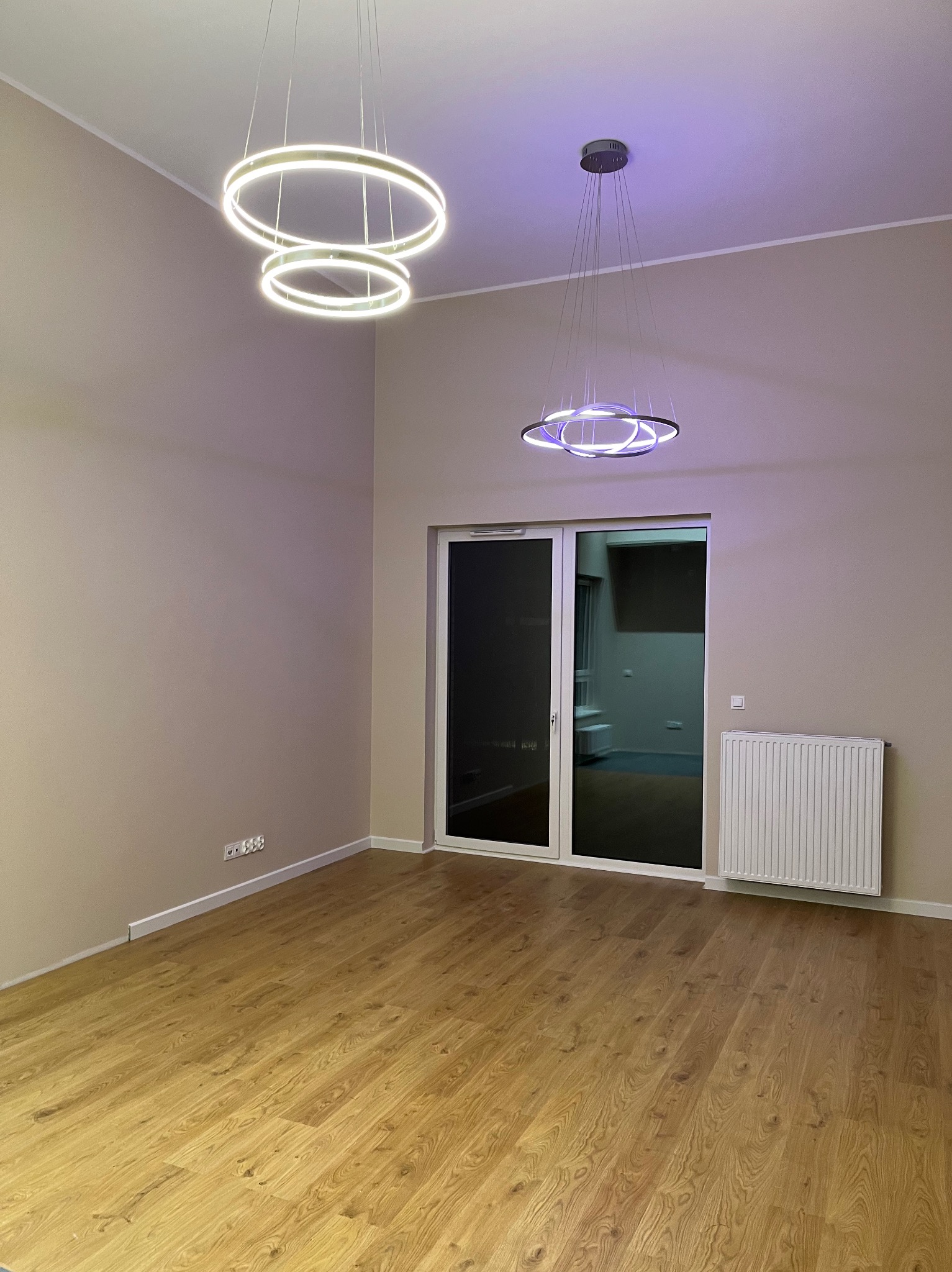 Pusty pokój z drewnianą podłogą, białymi ścianami, grzejnikiem i nowoczesnymi lampami wiszącymi w kształcie świecących okręgów.