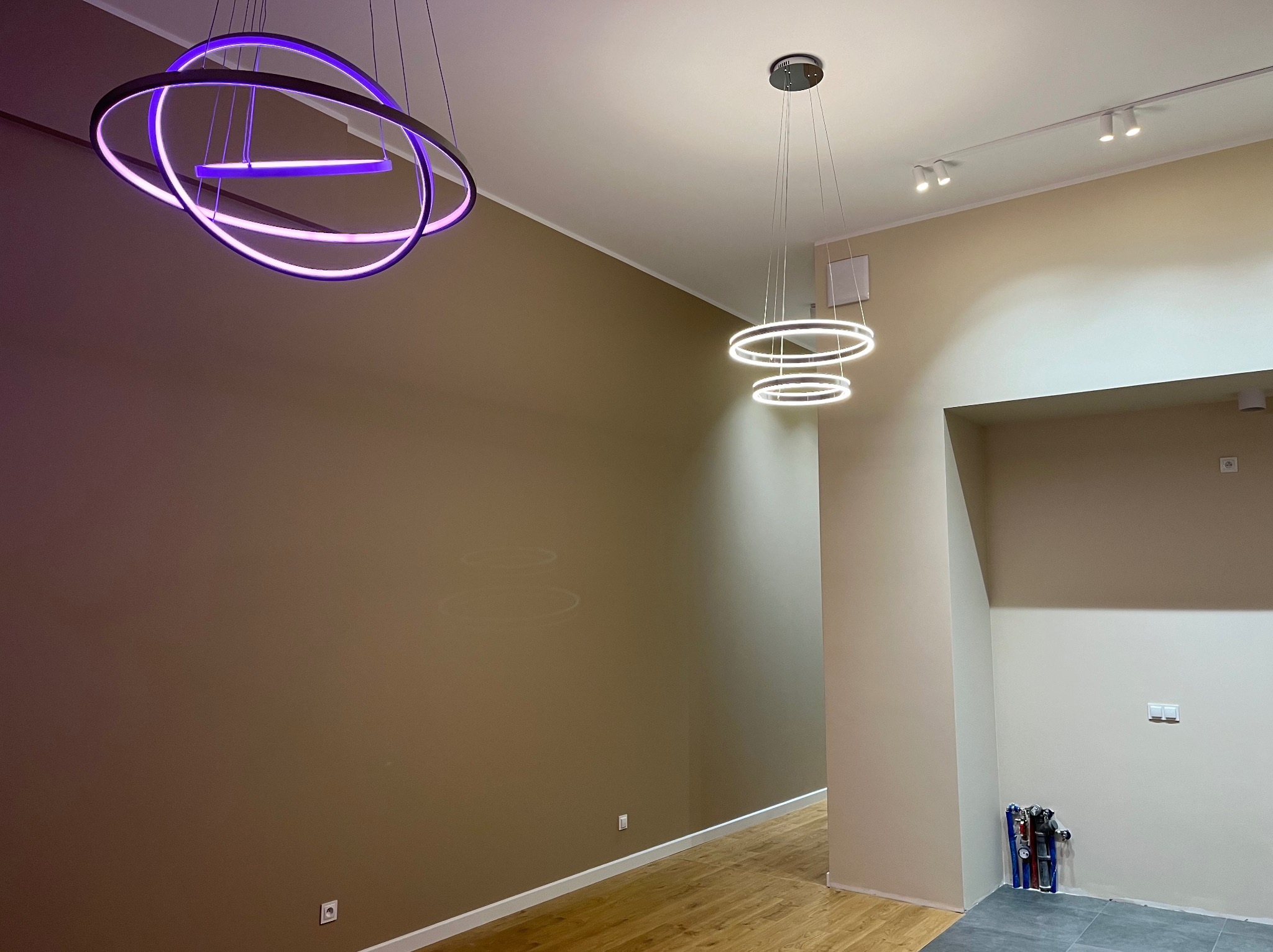Wnętrze z dwoma nowoczesnymi lampami wiszącymi: jedna z fioletowym światłem, druga z białym, oraz listwą z reflektorami na suficie. Ściany w odcieniach beżu i brązu, podłoga drewniana i szara...
