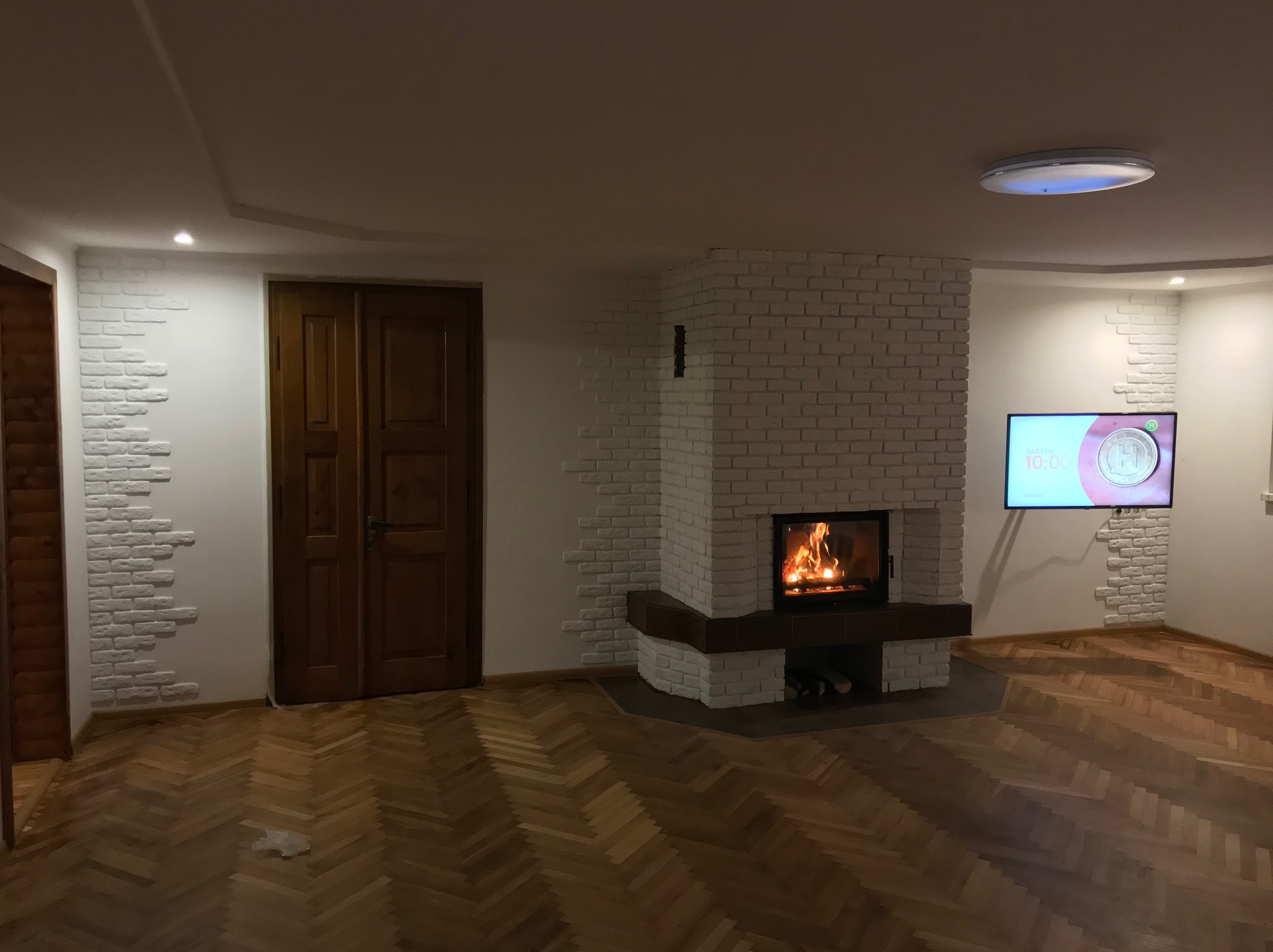 Salon z parkietem w jodełkę, kominkiem obłożonym białą cegłą i dekoracyjną ścianą z cegły, oświetlony lampą sufitową LED.