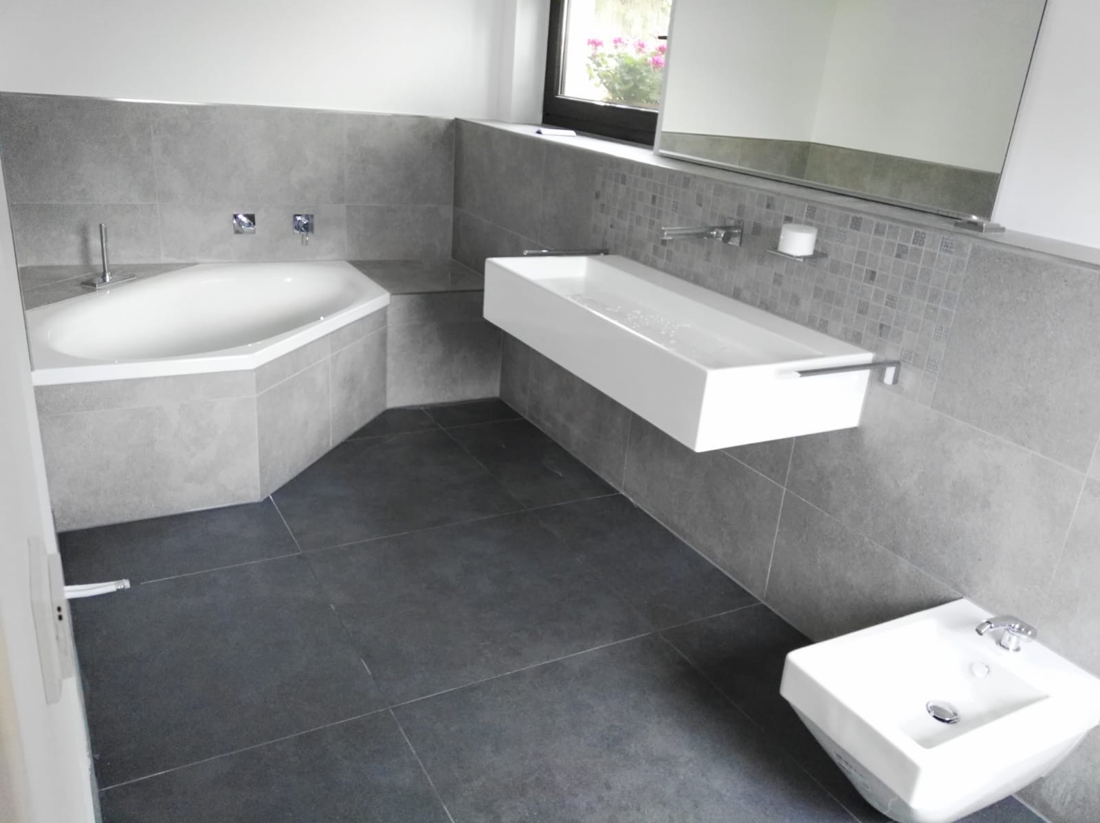 Nowoczesna łazienka w odcieniach szarości z narożną wanną, podwieszaną prostokątną umywalką i bidetem, wykończona płytkami ceramicznymi o różnych wzorach.