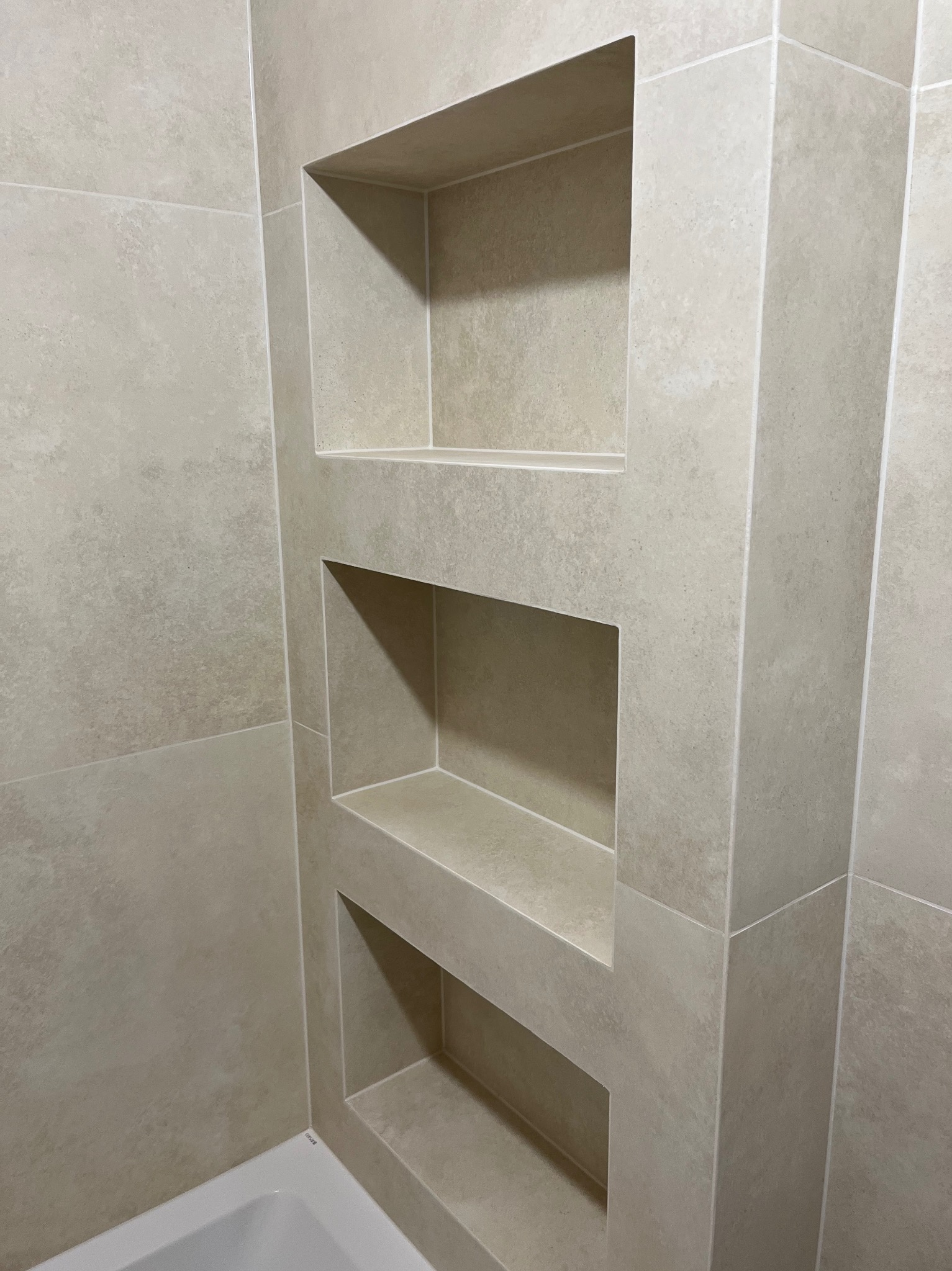 Beżowa ściana w łazience z trzema wnękami wyłożonymi płytkami. Minimalistyczny design, precyzyjne fugi, widoczny fragment białej wanny.