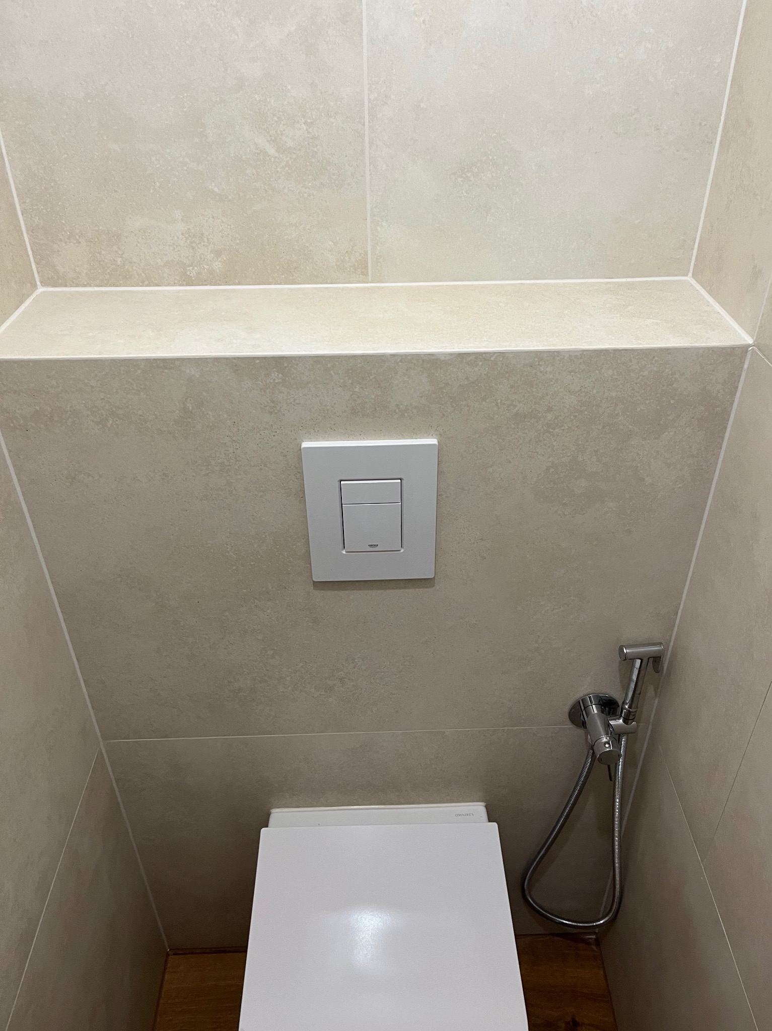 Minimalistyczna łazienka: beżowe płytki, biała toaleta podwieszana z przyciskiem spłukującym i bidetką. Drewniana podłoga w tle. Czyste linie, nowoczesny design.