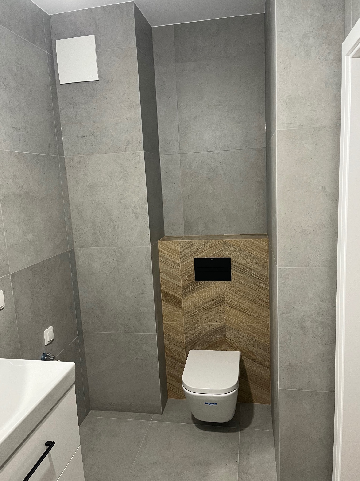 Nowoczesna toaleta podwieszana w szarej łazience z drewnopodobną mozaiką za zabudową. Minimalistyczny design, płytki imitujące beton, biała ceramika.