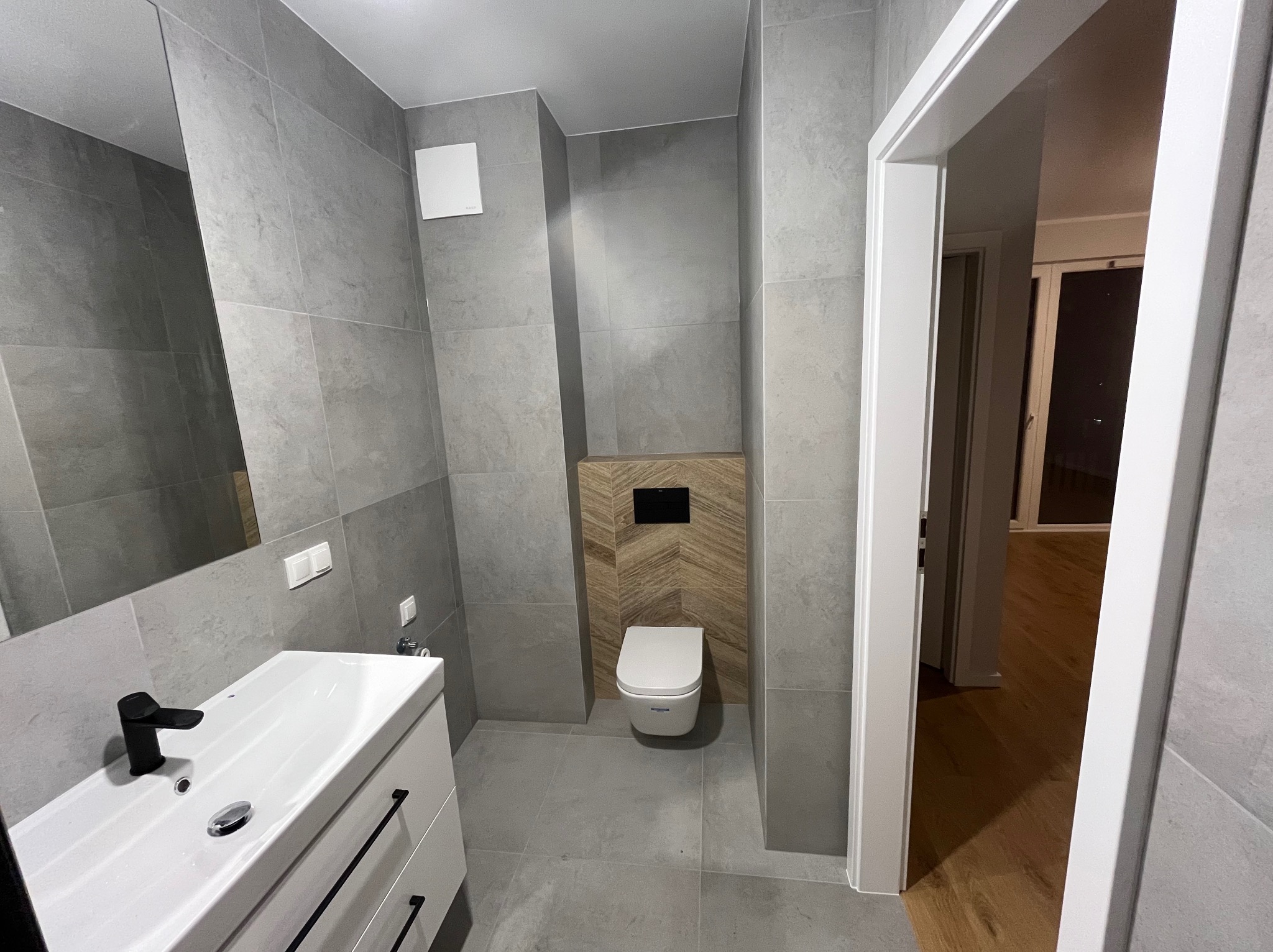 Nowoczesna łazienka w szarościach z akcentem drewna: umywalka z czarną armaturą, toaleta na tle drewnianej mozaiki, duże szare płytki i widok na inne pomieszczenie.