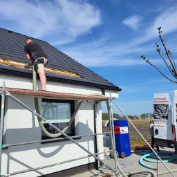 ADMAR Mariusz Pedda - Pracownik na rusztowaniu aplikuje materiał izolacyjny w szczelinę dachu domu jednorodzinnego, widoczna maszyna do wdmuchiwania izolacji oraz firmowy samochód dostawczy