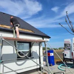 Pracownik na rusztowaniu aplikuje materiał izolacyjny w szczelinę dachu domu jednorodzinnego, widoczna maszyna do wdmuchiwania izolacji oraz firmowy samochód dostawczy