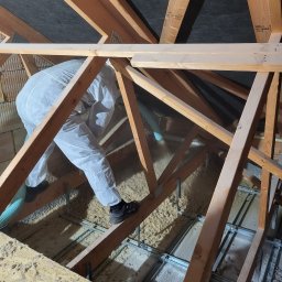 ADMAR Mariusz Pedda - Pracownik w kombinezonie ochronnym aplikuje materiał izolacyjny na poddaszu za pomocą węża, widoczne drewniane elementy konstrukcji dachu oraz metalowy stelaż pod podłogą.